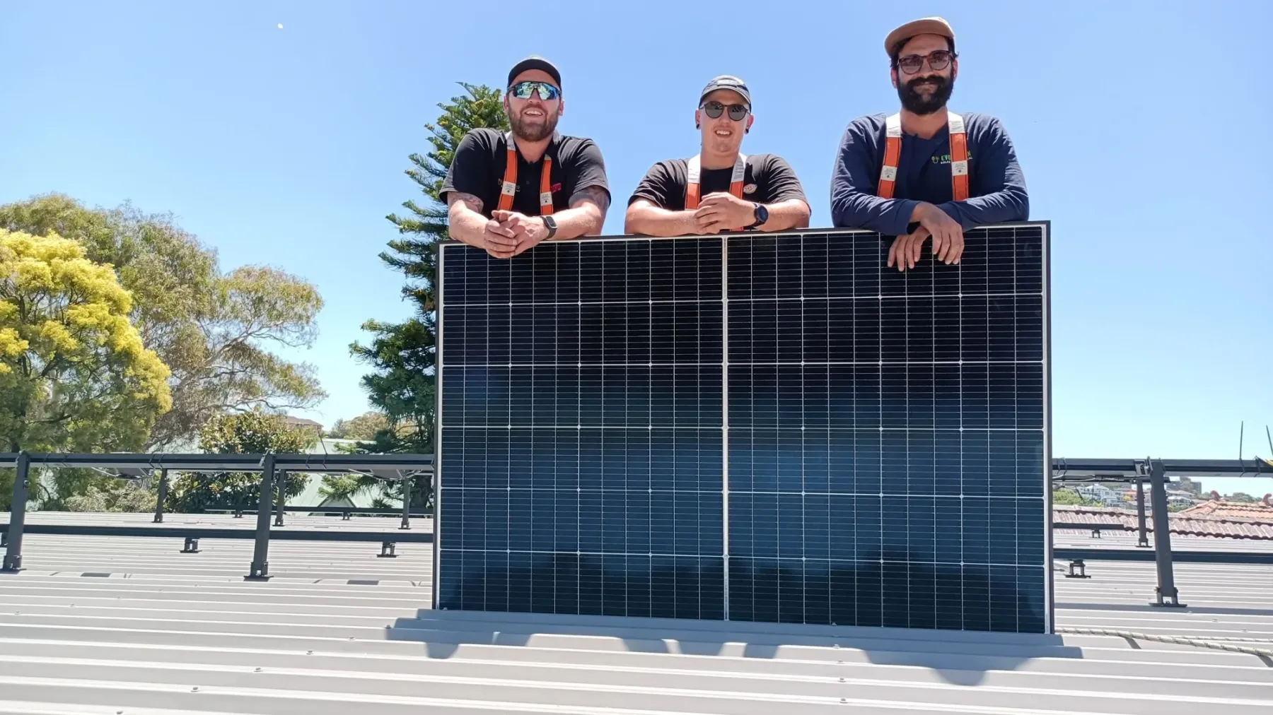 Three solar installers on a metal roof leaning on a large solar panel
