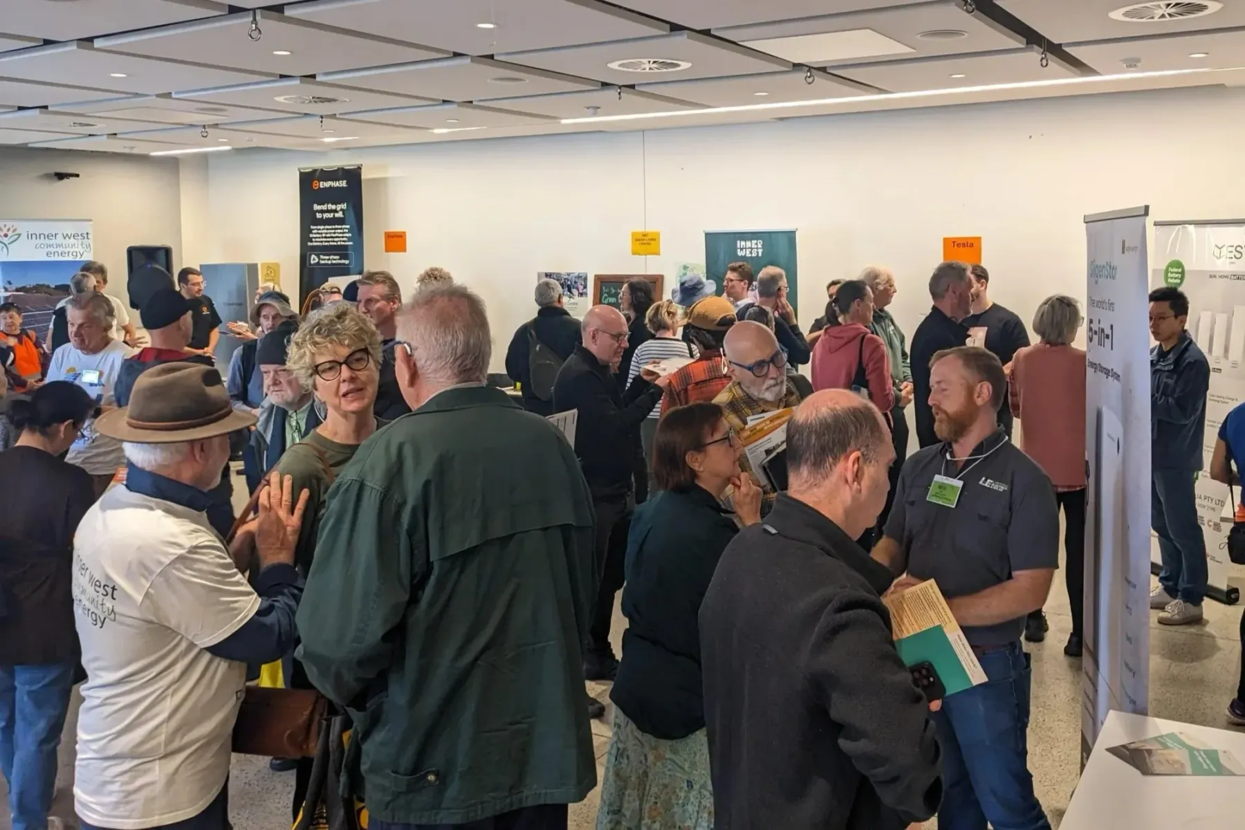 In a large room, a crowd of people mingles with stallholders. Various signage is on display.
